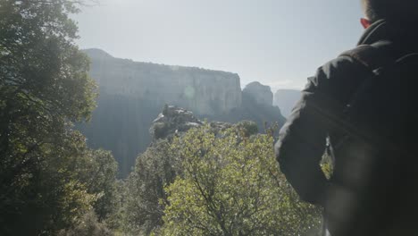 man wearing hat and jacket walks down trail with trees in forest, stops, takes hat off, observes spectacular scenic landscape view of mountain range and lake, pan behind