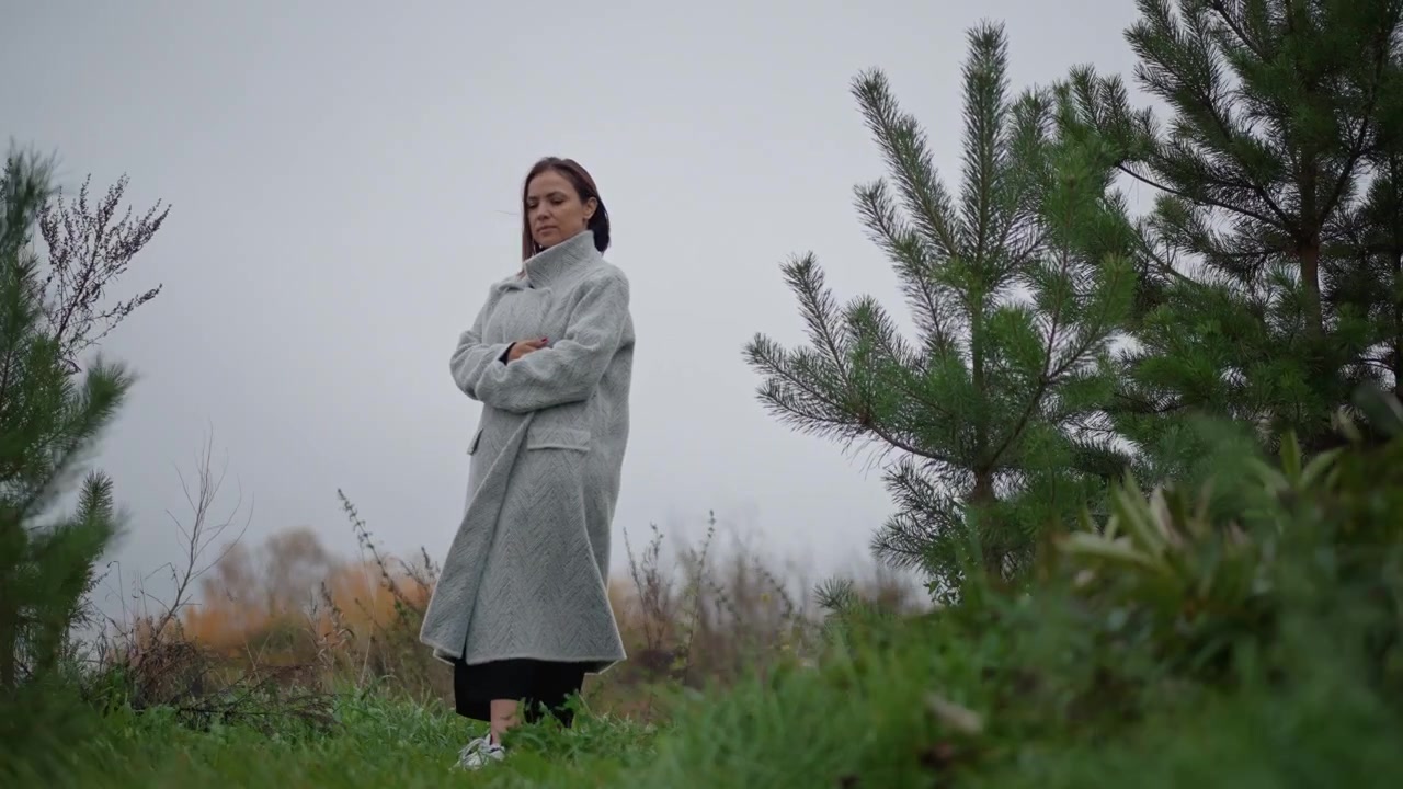 Giovane donna con un lungo cappotto chiaro e pantaloni scuri sta su una collina erbosa con le braccia incrociate e un'espressione neutra, vista da un'angolazione bassa sotto una luce soffusa e nuvolosa con una leggera brezza e una calma solitudine.