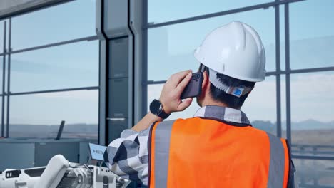 vista cercana de atrás de un ingeniero asiático con casco de seguridad hablando en su teléfono inteligente mientras está de pie en una fábrica de alta tecnología, analizando el concepto de máquina robótica