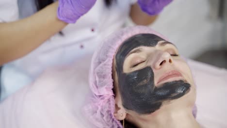 A doctor-cosmetologist applies a black mask with a brush to cleanse and moisturize the skin of the face in the beauty center. Close-up.