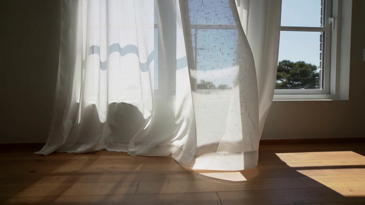 Billowing sheer curtains drifting on wood floor at open window, pushed by breeze with sun bands