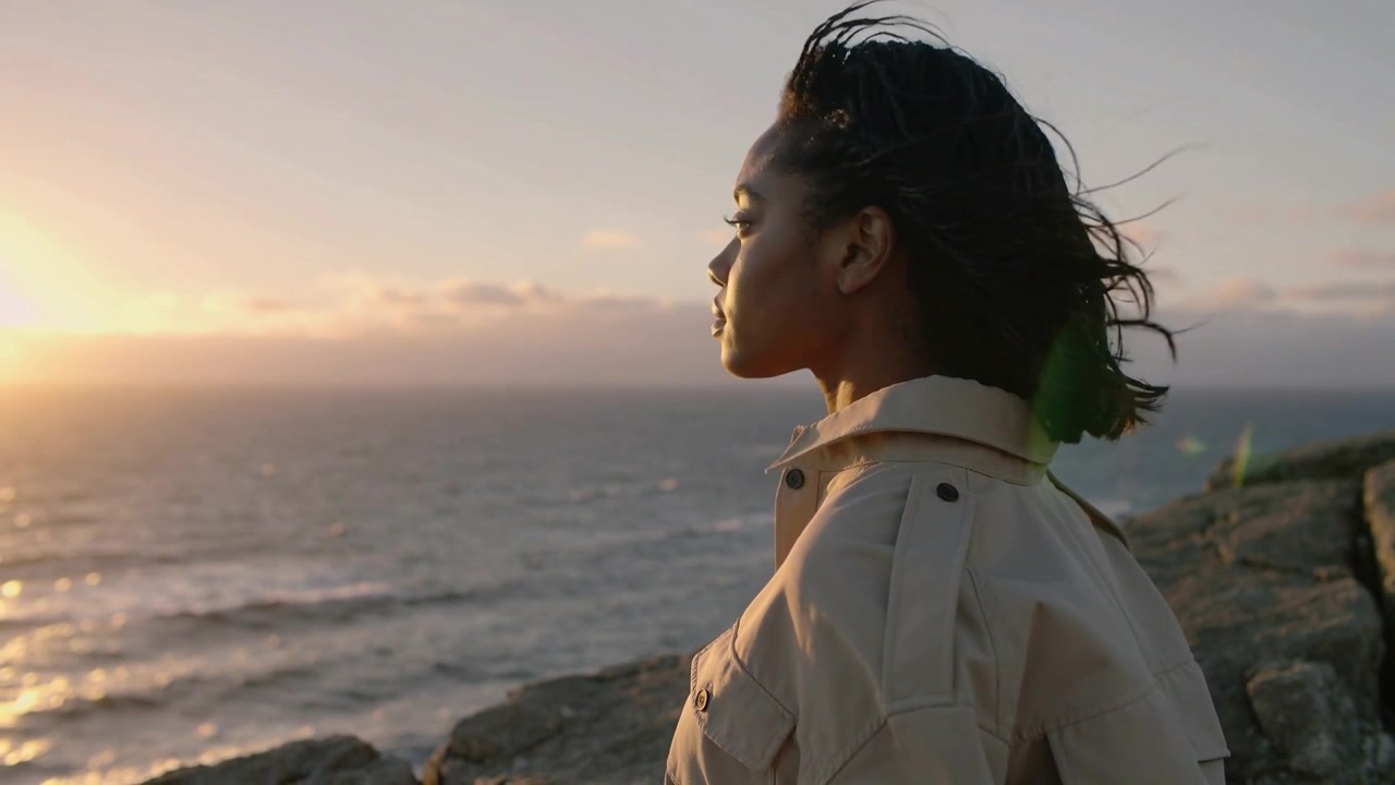 Gazing woman responding to sun lowering, turning to ocean on rock cliff, beige jacket, copy space