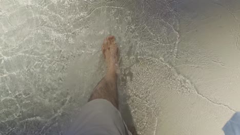 pov of a caucasian male walking in crystal-clear shallow waves on soft white sand. wearing beige swim trunks, his feet move through the sunlit water, with droplets sparkling in the sunlight.