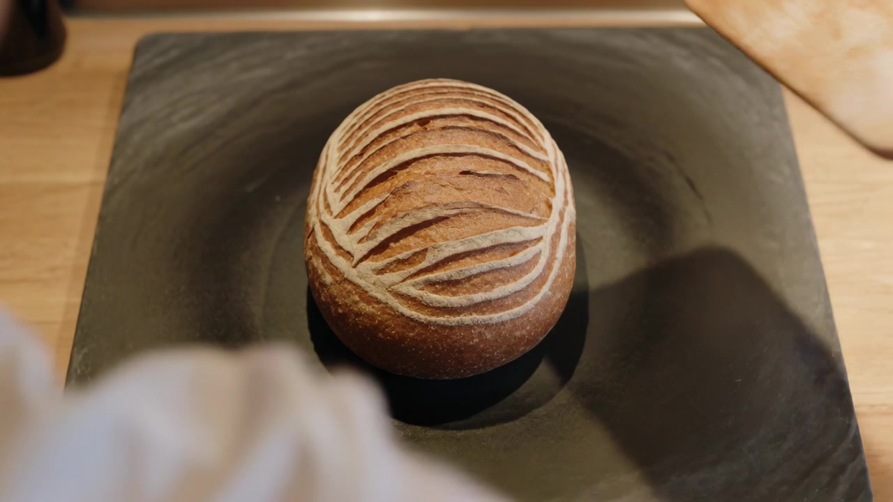 Entering with peel, baker wearing apron placing scored loaf onto stone on counter, for baking