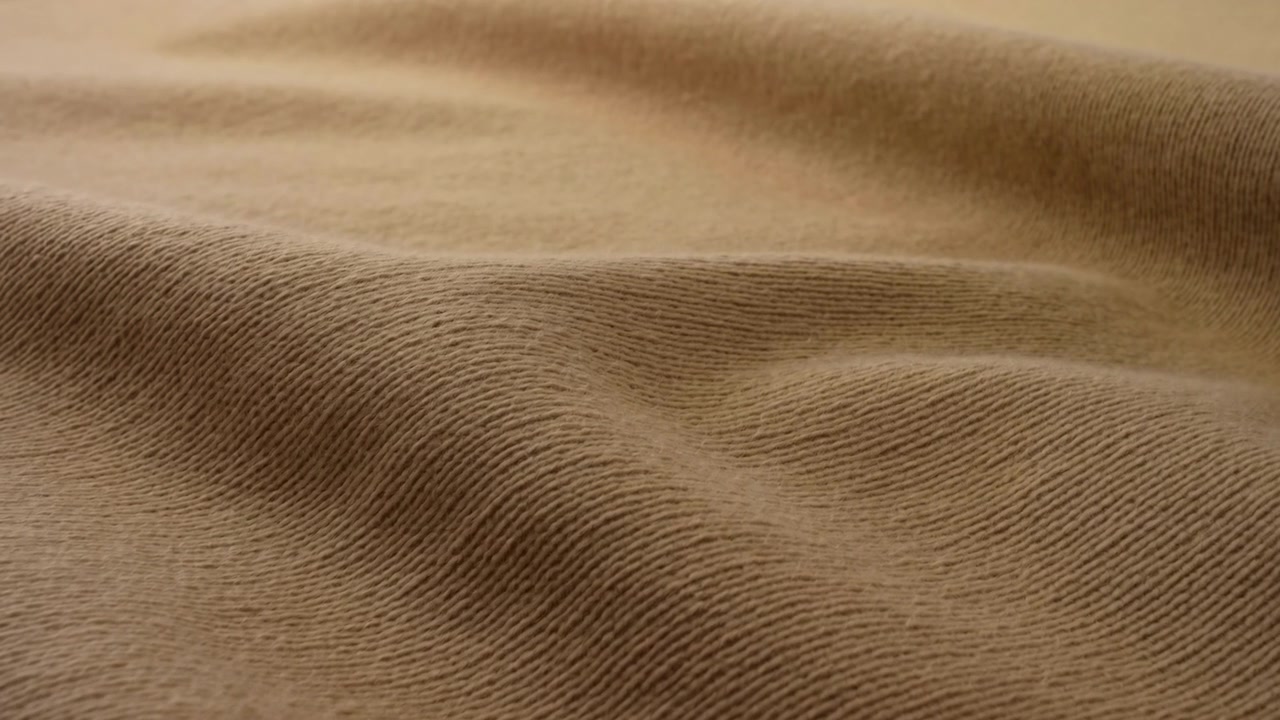 Nærbildebevegelse av beige ribbestrikket stoff som viser en forsiktig legging og omforming av overflaten til grunne åser og daler, mykt, diffust lys fra øvre venstre side skaper subtile gradienter og lav kontrast