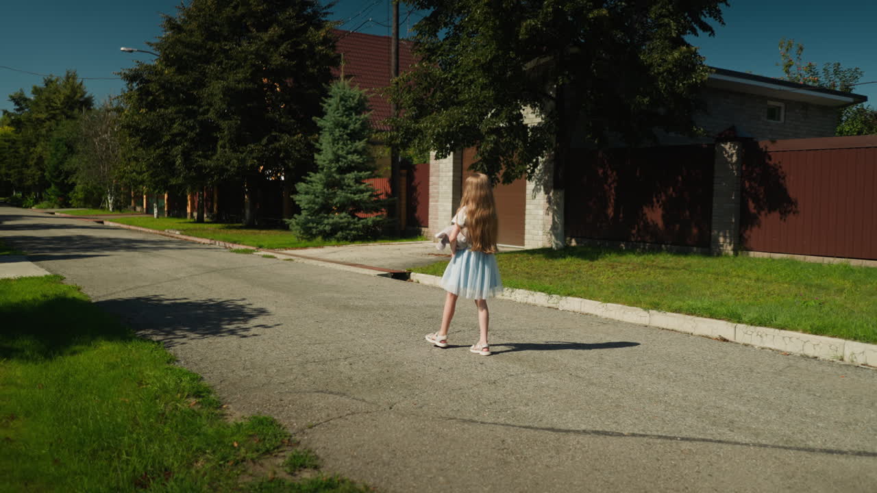 Little girl in light blue dress walking alone on quiet residential street during sunny day, holding soft teddy bear, surrounded by green grass, trees, fences, and houses with shadows on pavement