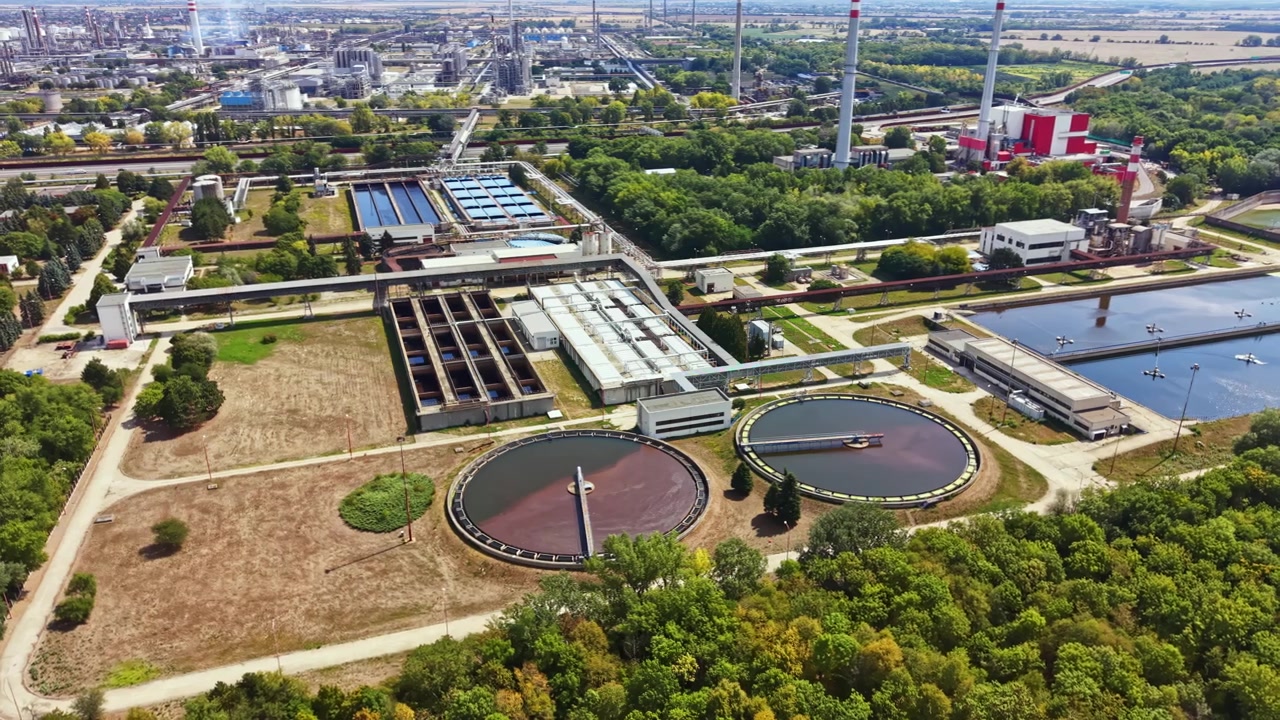Factory and water treatment facility located near a green area with trees, showing various large tanks on a sunny day