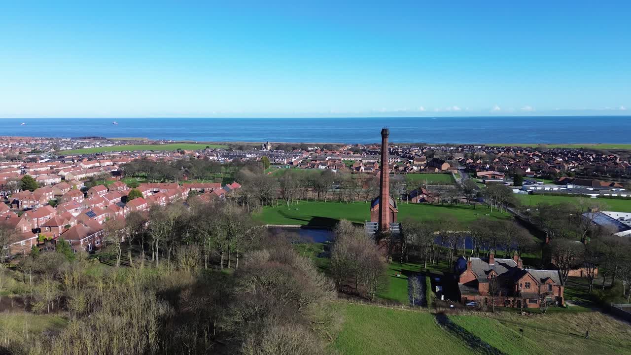 Aproximação aérea do Museu Ryhope Engines: um voo de drone sobre campos verdes em direção à alta chaminé de tijolos, com os subúrbios de Sunderland e a costa do Mar do Norte visíveis em plena luz do dia.