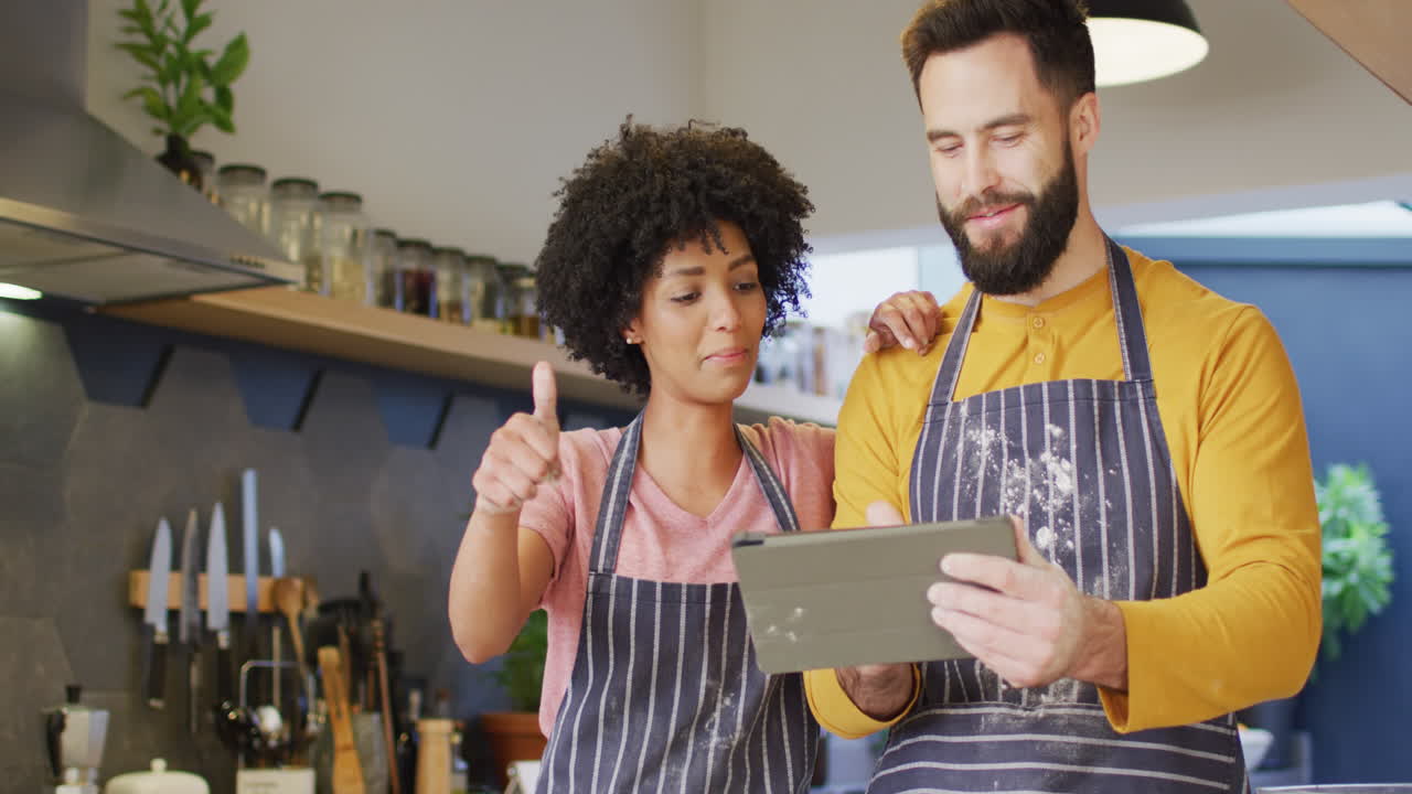 video de una feliz pareja diversa en delantales usando una tableta, horneando juntos en la cocina, con espacio para copiar