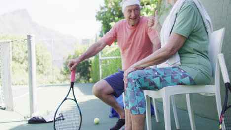 happy senior caucasian couple taking a break, talking at sunny outdoor tennis court, slow motion