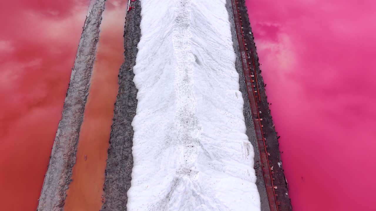 Top down view of a long pile of processed white salt located on a grey path between pink and orange tinted water. Close up aerial shot of white salt ridge between ponds