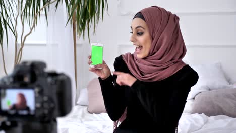 young beautiful indian girl in hijab recommends an application on a smartphone, smiling, talking at the camera, chroma key, green screen concept. 50 fps
