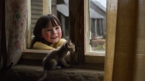 a young girl smiles at a kitten through an old window