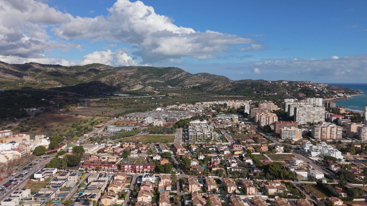 Benicassim,Castellon, Spain slow forward drone footage towards the Desert de les Palmes mountains and the coast flying over the town on bright sunny day with blue but cloudy sky