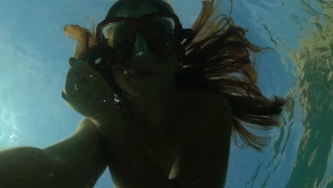 selfie bajo el agua de una joven pelirroja con máscara de buceo haciendo burbujas de aire mientras flota en agua de mar transparente