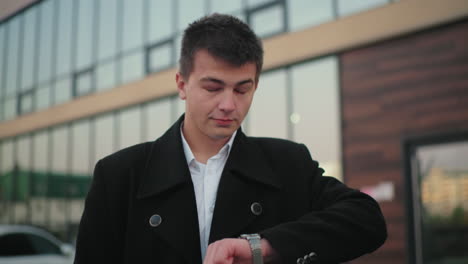 real estate mogul in black coat checking time with confident look, background featuring modern glass building, professional, successful businessman exuding confidence in urban environment