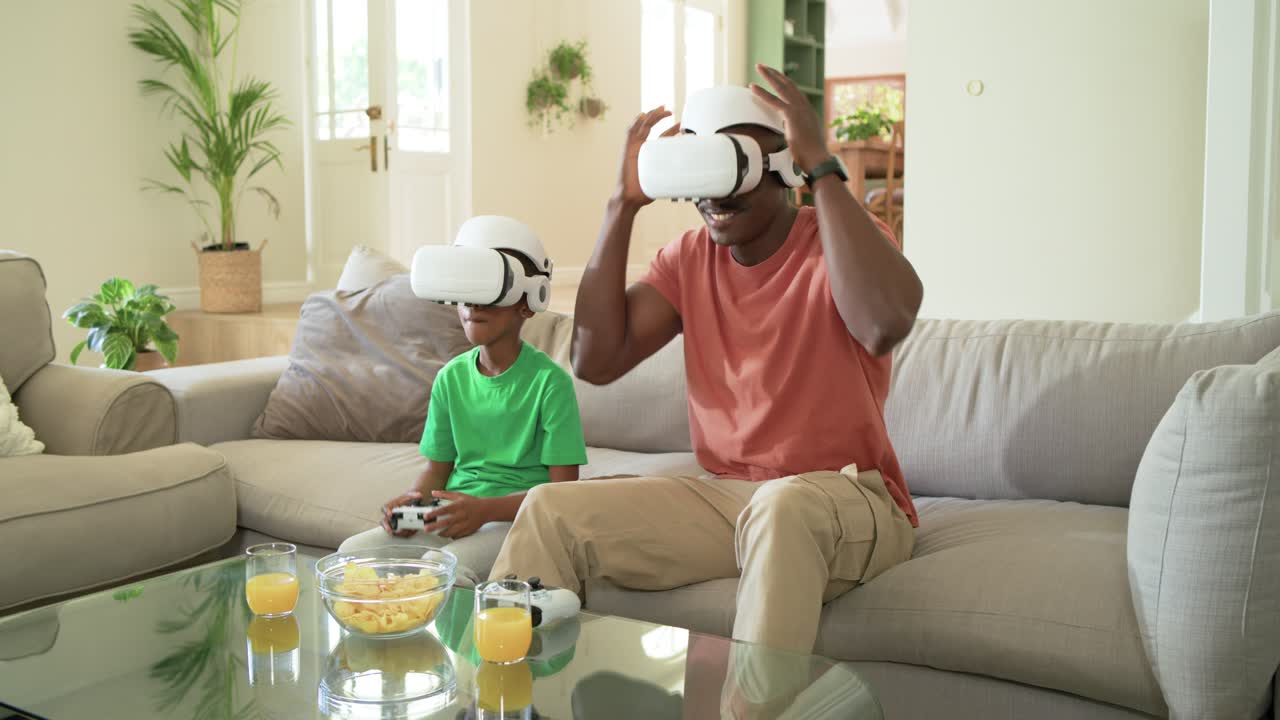 Padre e figlio afroamericani che sistemano un visore VR bianco, passano il controller e giocano sul divano