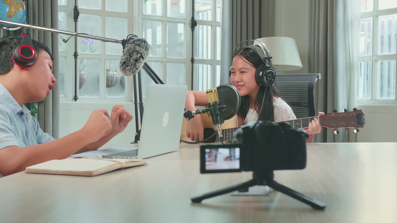 Asian Kid Girl In Headphones Playing The Guitar And Singing Into Microphone While Recording Podcast With Boy Host In Studio