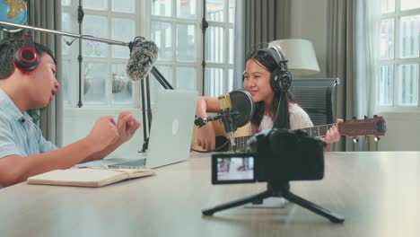 asian kid girl in headphones playing the guitar and singing into microphone while recording podcast with boy host in studio