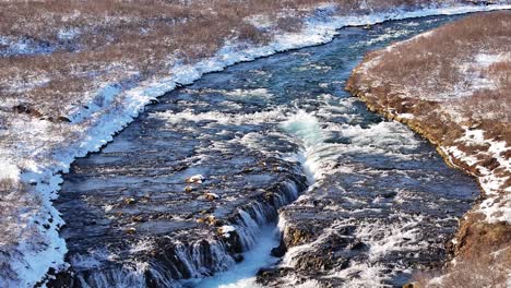 snowy landscape with icy blue river cascading over brúarfoss waterfall in iceland