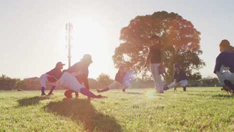 Diverse-group-of-female-baseball-players-and-coach-on-sunny-pitch,-squatting-and-stretching-legs