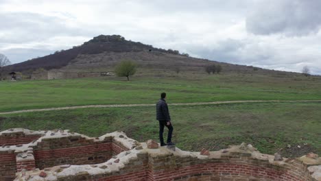 Man-walking-on-a-fallen-brick-wall-in-a-remote-location-in-an-ancient-city-in-Bulgaria,-with-mountains-in-the-background