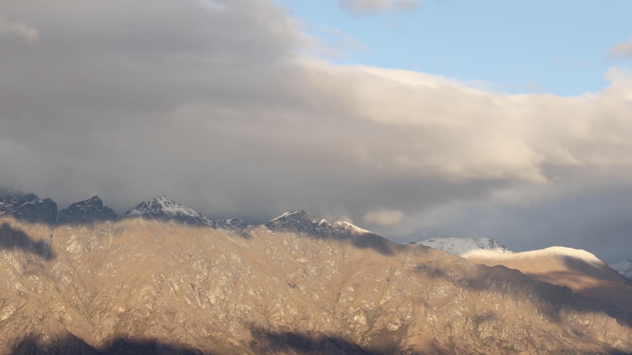 Clouds move swiftly over a rugged mountain range, casting dynamic shadows and highlighting the landscape's natural beauty