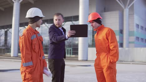 Inversionista-Del-Proyecto-Con-Traje-Negro-Examinando-El-Objeto-De-Construcción-Con-Trabajadores-De-La-Construcción-Con-Uniforme-Naranja-Y-Cascos.-Están-Revisando-Los-Dibujos-Usando-La-Tableta.