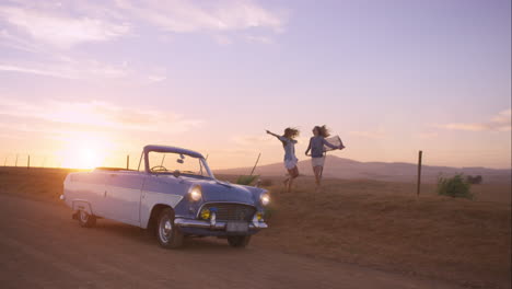 Girl-friends-dancing-at-sunset-on-road-trip-with-vintage-car
