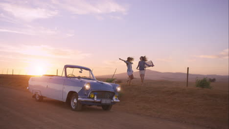 girl friends dancing at sunset on road trip with vintage car