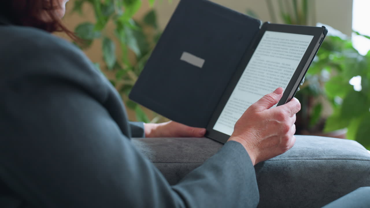 Middle-aged woman reading digital book on e-reader while relaxing on gray couch surrounded by indoor plants in cozy living room environment during peaceful afternoon moment of solitude