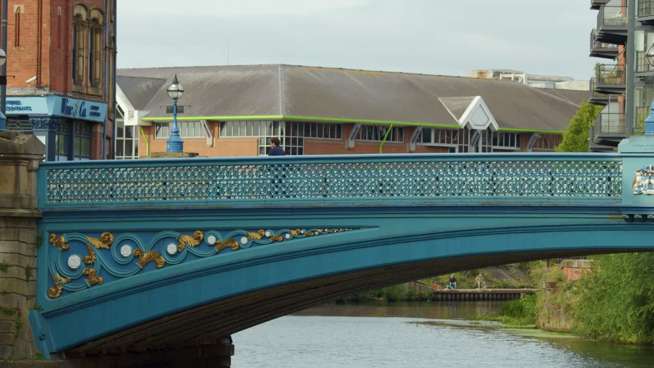 Two people walk across a decorative blue bridge over calm river, daylight, static camera