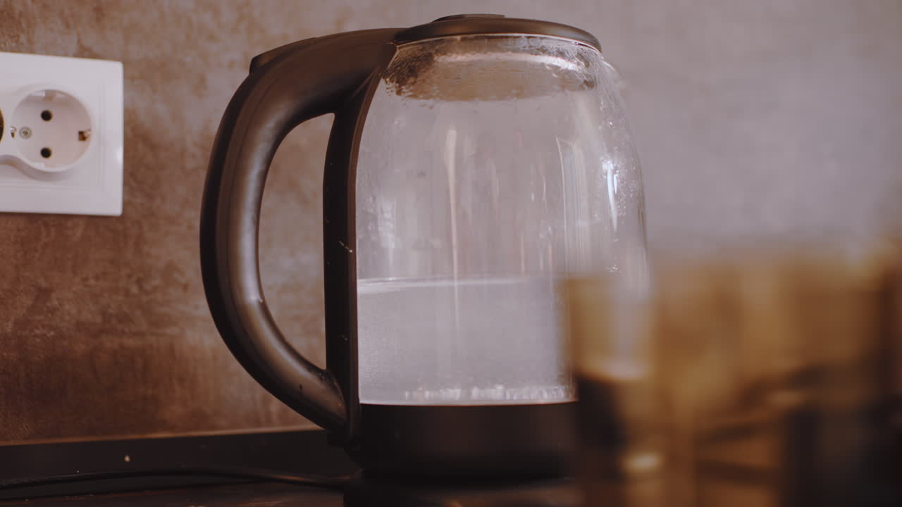 Primer plano de un hervidor eléctrico transparente hirviendo agua con vapor formándose en la superficie, mientras que una taza de cristal se sitúa en primer plano sobre la encimera de la cocina, creando una preparación de bebida caliente.