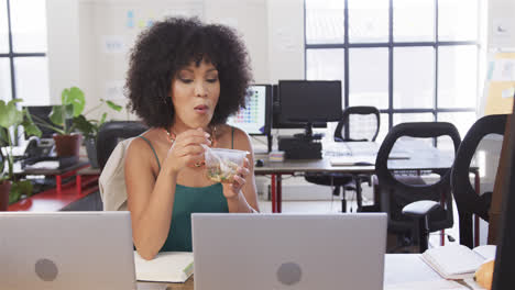 feliz mujer de negocios casual biracial sentada en el escritorio, comiendo y mirando la computadora portátil, cámara lenta