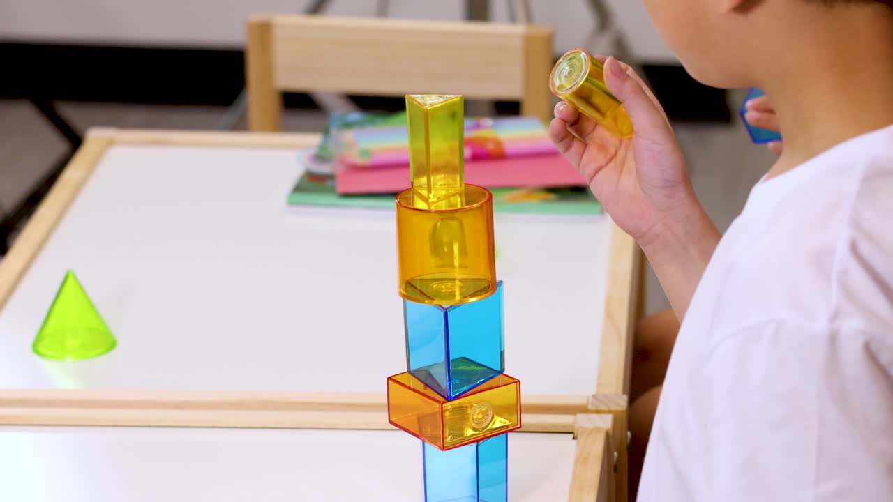 Anak kecil dengan hati-hati melepaskan balok-balok transparan dari menara tinggi di suasana ruang kelas yang terang benderang