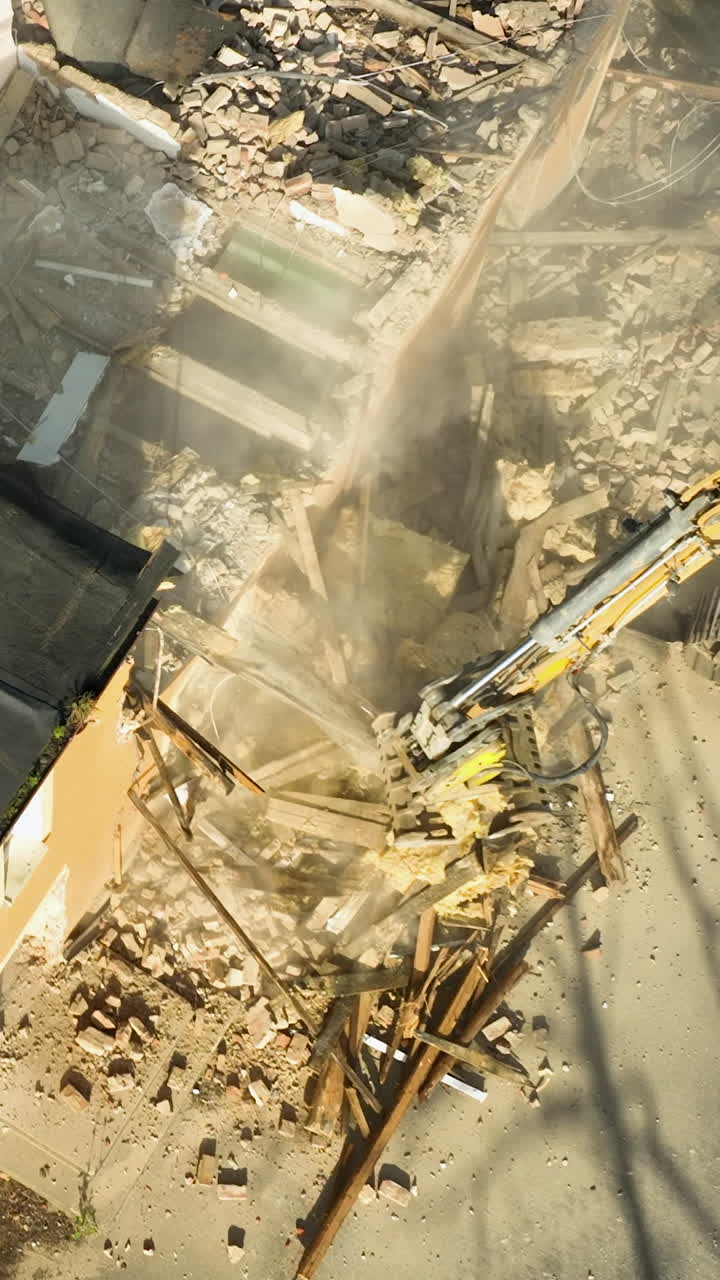 Un plan cinématographique en plongée d'une pelleteuse jaune démolissant une structure, créant de la poussière et des débris. Utile pour les projets de construction, de rénovation urbaine et de développement industriel.