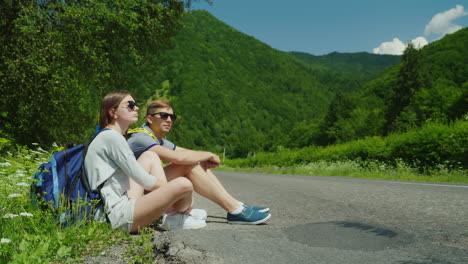 a couple of tired tourists with backpacks rest on the roadside in a picturesque place in the backgro