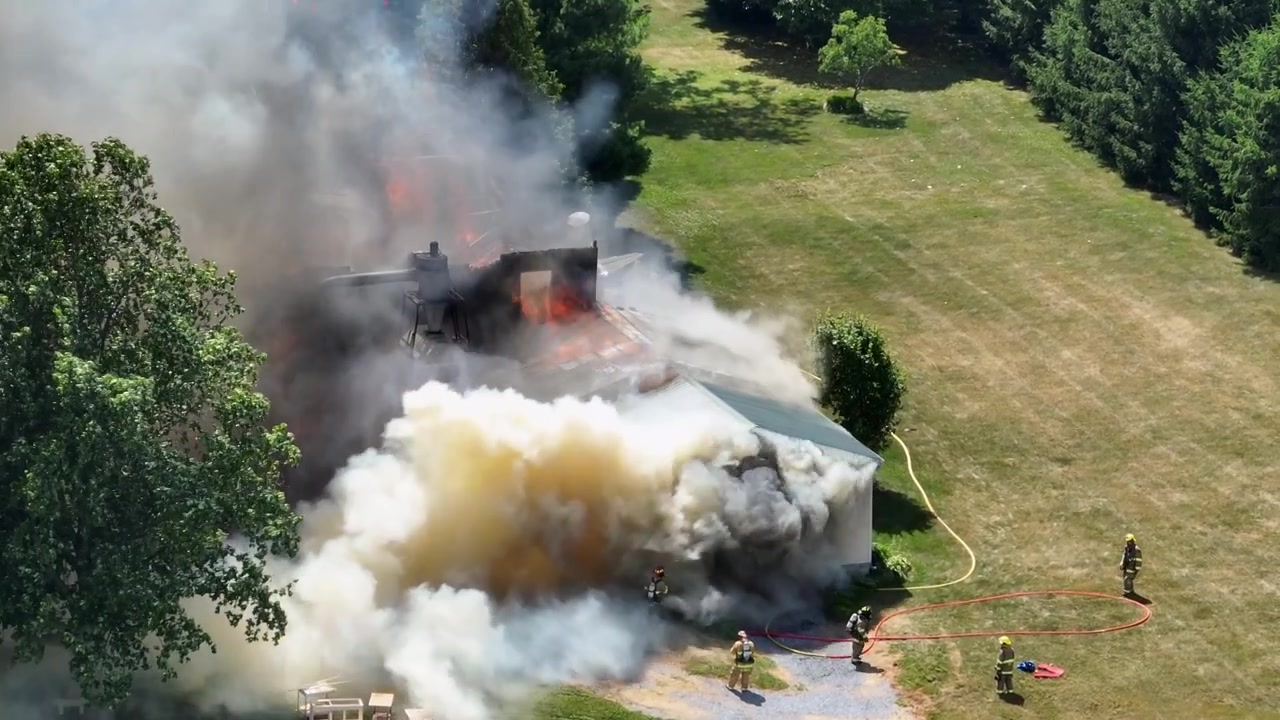 Vigili del fuoco in abiti protettivi che spruzzano acqua con la manichetta su una casa in fiamme in un'area rurale. Giornata di sole su terreni agricoli di una città americana. Densi fumi tossici che salgono nel cielo. Ripresa aerea dall'alto.