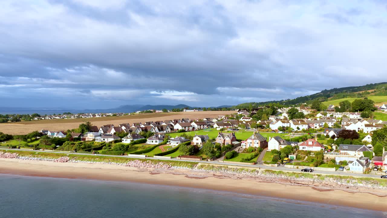 Drone pans above Rosemarkie village, beachfront houses, green hills, and sandy shoreline in daylight