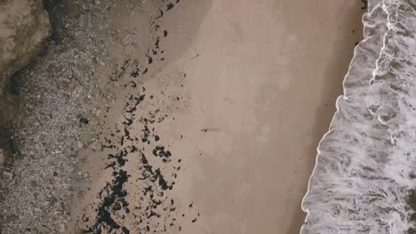 aerial birds eye view drone footage of a man on an empty beach with big ocean sea waves crashing onto the shoreline