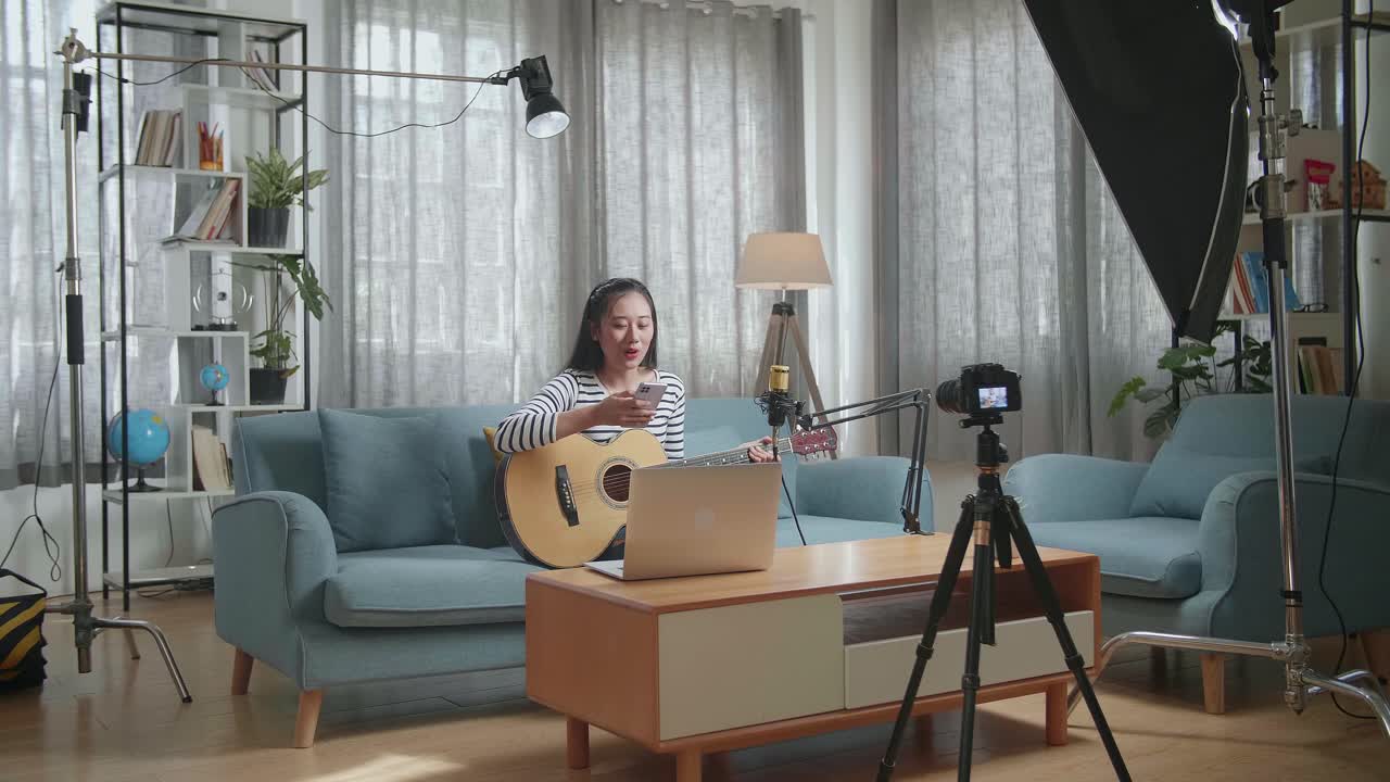 노트북을 가진 아시아 여성이 스마트폰에서 글을 읽고 라이브 스트리밍을 하는 동안 카메라에 대화하고 집에서 기타를 연주합니다.