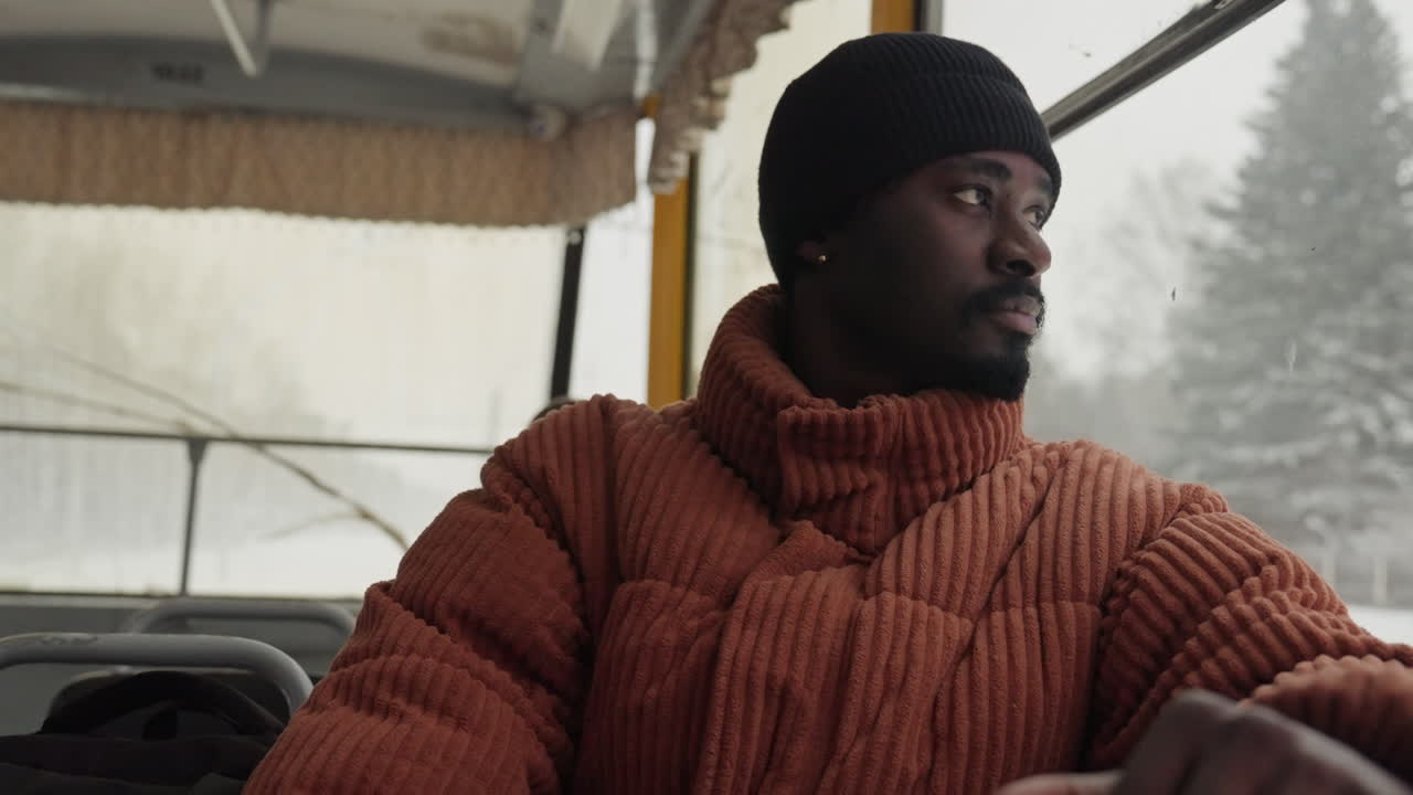 Man on bus by snowy window gazing into distance wearing chunky knit jacket and beanie, soft ambient light, muted color palette, quiet reflective mood, urban winter landscape outside, seated passenger