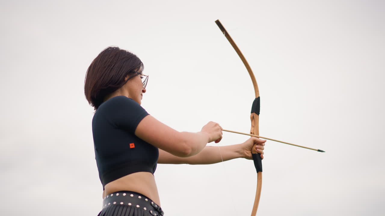 Bogenschützin im Freien, die einen Bogen spannt, stabile Haltung und konzentriertes Zielen unter bewölktem Himmel, Lederköcher an der Hüfte, Recurve-Bogen mit eingelegtem Pfeil zurückgezogen, angespannter Moment vor dem Lösen, der Ruhe vermittelt