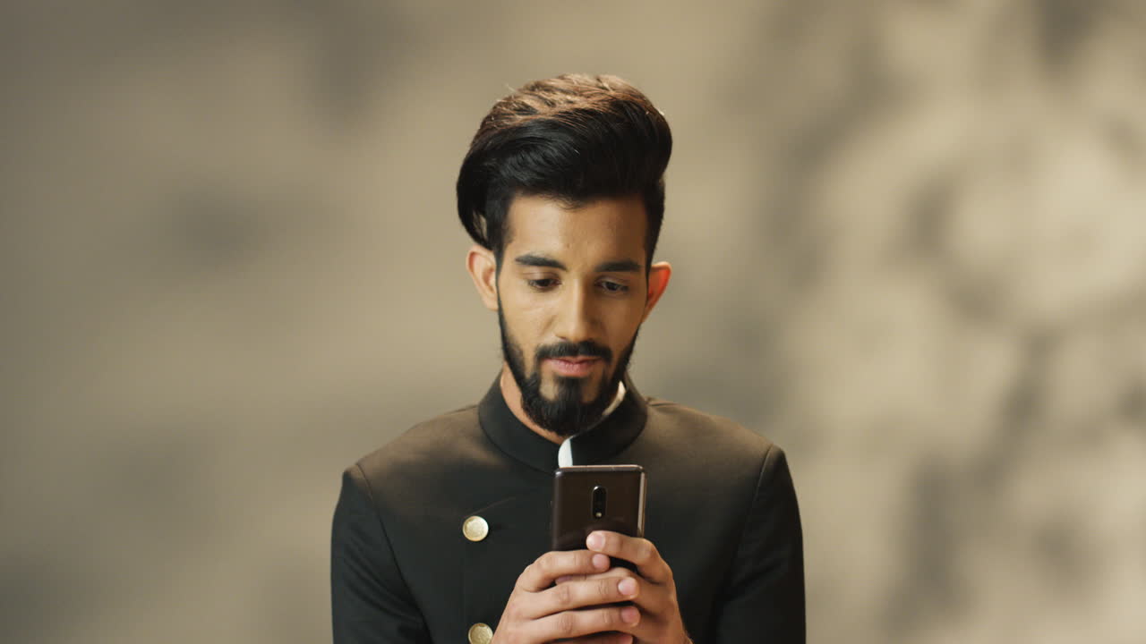 Giovane uomo indiano allegro con la barba e in giacca nera che scrive messaggi sullo smartphone