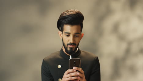 joven indio alegre con barba y chaqueta negra enviando mensajes de texto en el teléfono inteligente