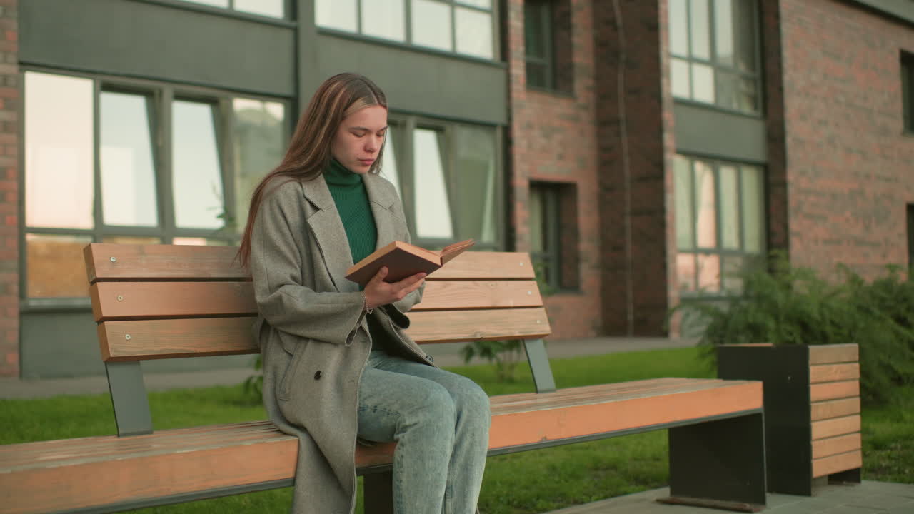 Woman seated on wooden bench outdoors reading book then lifting head thoughtfully, wearing gray coat and green turtleneck with residential building background
