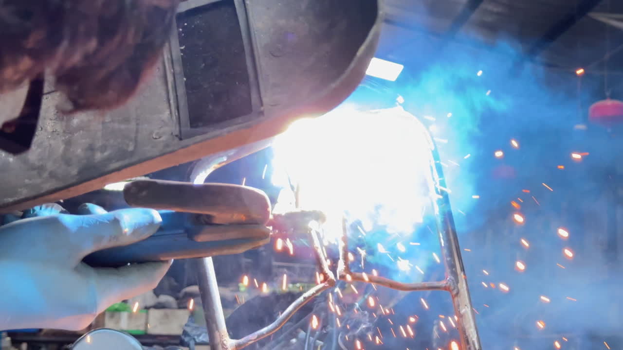 Low-angle close-up of a person welding a motorcycle frame, surrounded by bikes under a workshop roof.