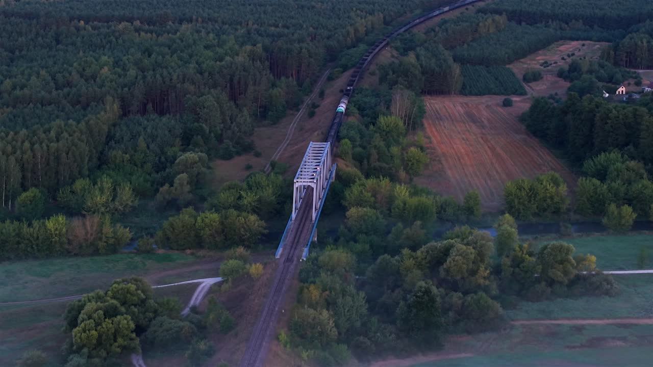 Aerial establishing view, freight train crossing an old steel railway bridge over the Nida River in Poland, surrounded by forest and fields in soft morning