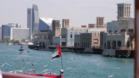 many seagulls are flying behind a boat on the sea of dubai, in the background is the huge skyline of dubai