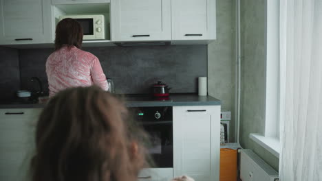 woman in pink velvet robe stands at kitchen counter preparing food while girl with ponytail watches from behind in bright room, evoking quiet domestic moment during morning breakfast routine